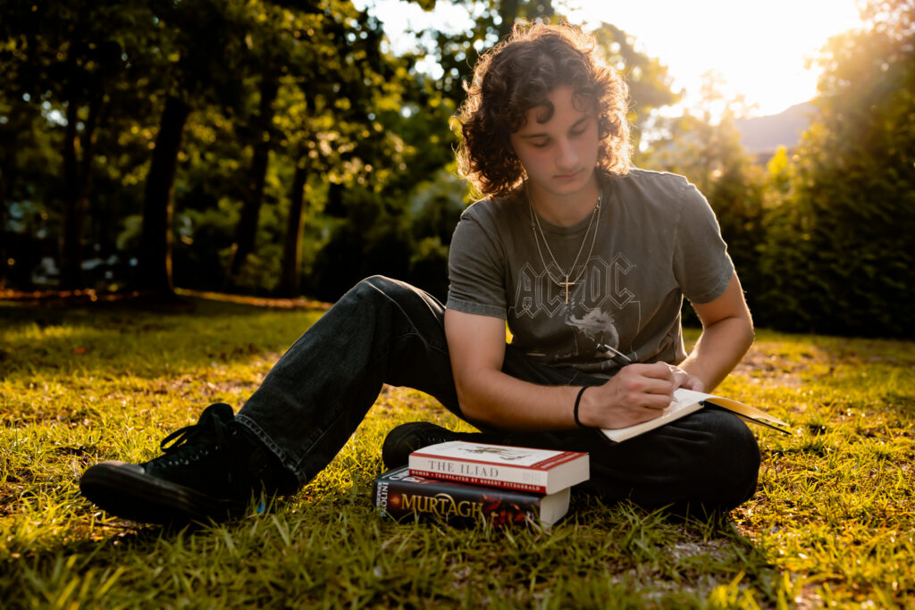 color photo of a young man sitting in the grass outside writing in a journal
