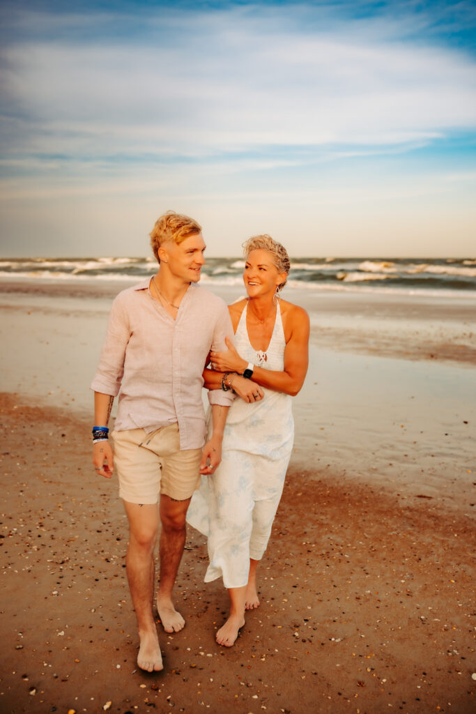 color photo of a mom and her adult son laughing and walking on the beach.