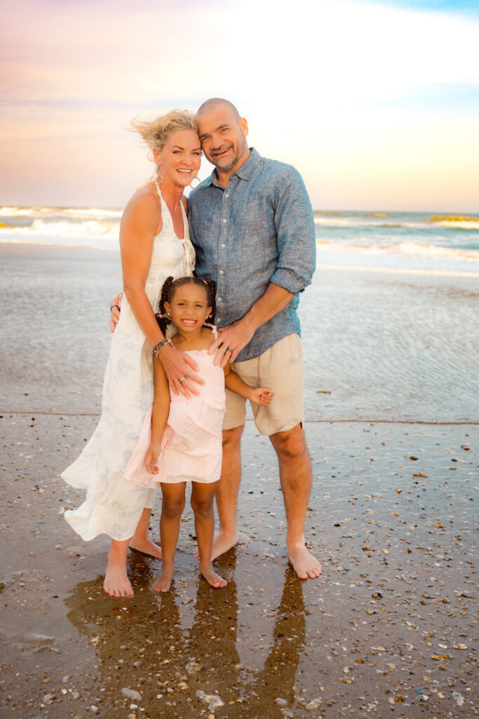 Color photo of grandparents standing with their granddaughter on the beach smiling.